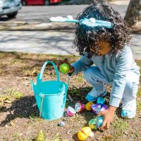 Little girl with little bucket picking up plastic easter eggs outside taking part in one of these 5 Outdoor Easter Egg Hunts