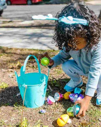Little girl with little bucket picking up plastic easter eggs outside taking part in one of these 5 Outdoor Easter Egg Hunts